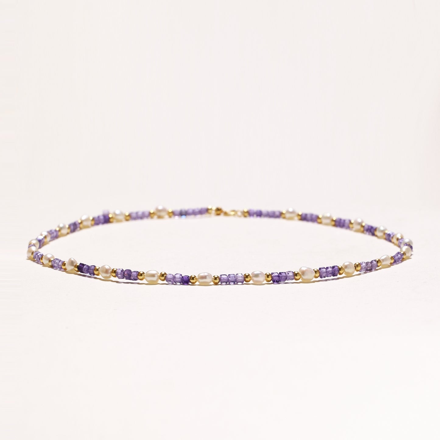 Delicate beaded necklace with alternating purple zirconia stones, white freshwater pearls, and gold-plated spacers on cream background.