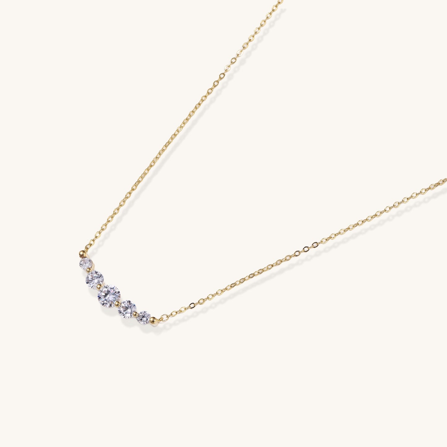 Delicate 14K gold plated necklace with curved arc of sparkling zirconia stones on cream background.