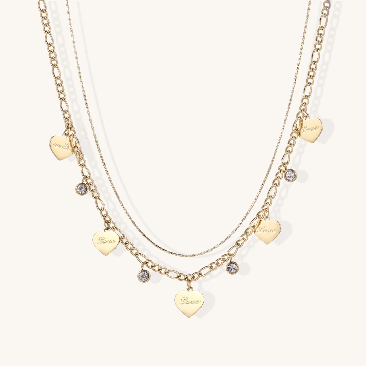 Two layered gold-plated necklaces with heart pendants engraved 'Love' and sparkling crystal charms on white background.