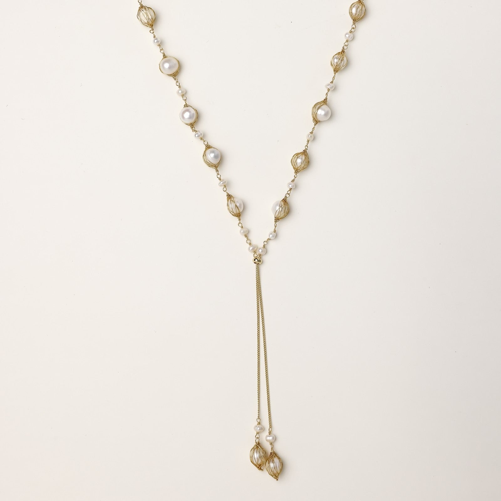 Long gold-plated lariat necklace with wire-wrapped freshwater pearls and chain tassel detail on cream background.
