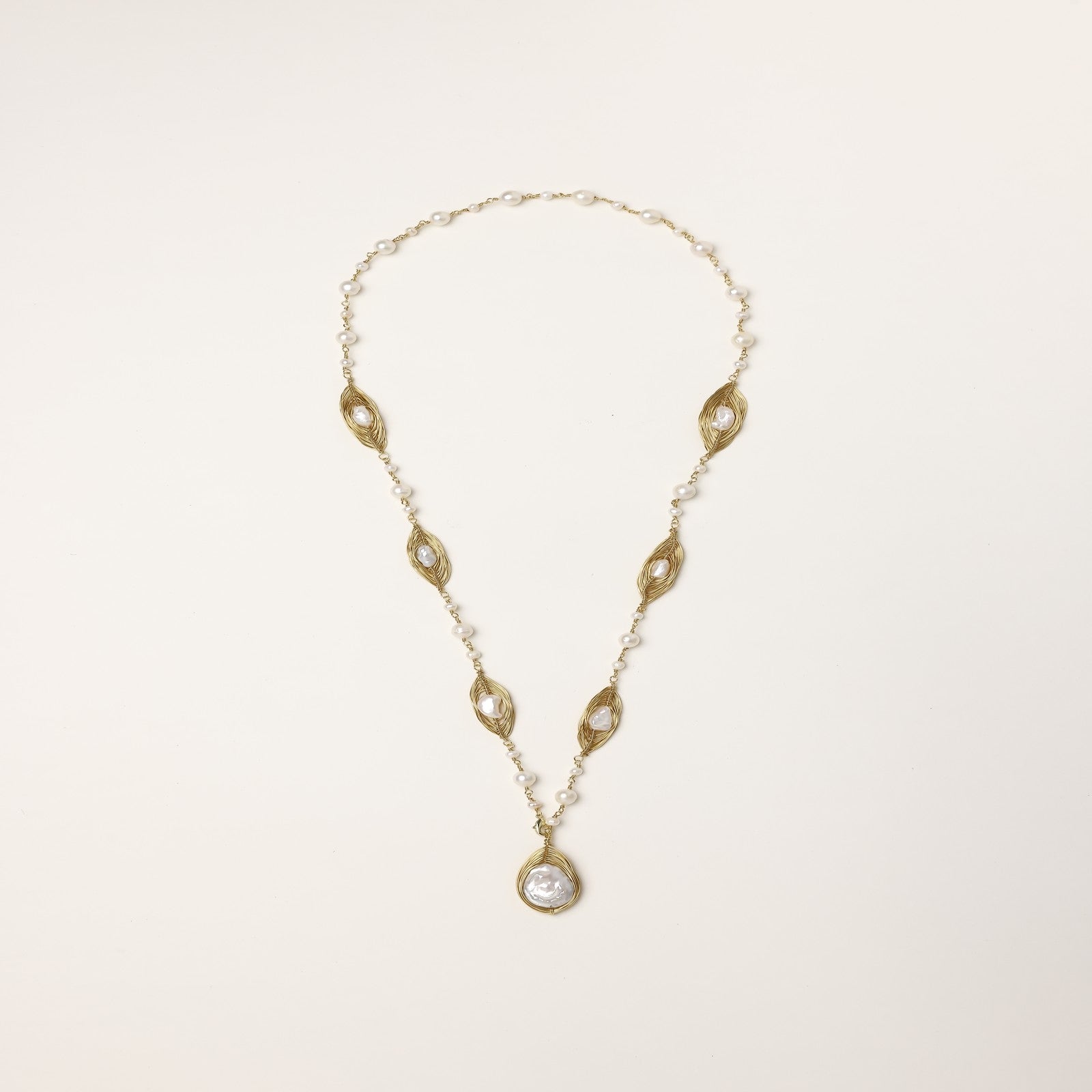 Long gold-plated necklace with eye-shaped elements, freshwater pearls, and baroque pearl pendant on neutral background.