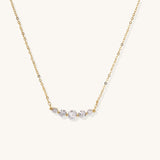 Delicate 14K gold plated necklace with curved arc of sparkling zirconia stones on cream background.