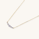 Delicate 14K gold plated necklace with curved arc of sparkling zirconia stones on cream background.