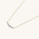 Delicate 14K gold plated necklace with curved arc of sparkling zirconia stones on cream background.