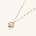 Gold-plated pendant necklace with pink enamel background and white freshwater pearl floral design on neutral background.