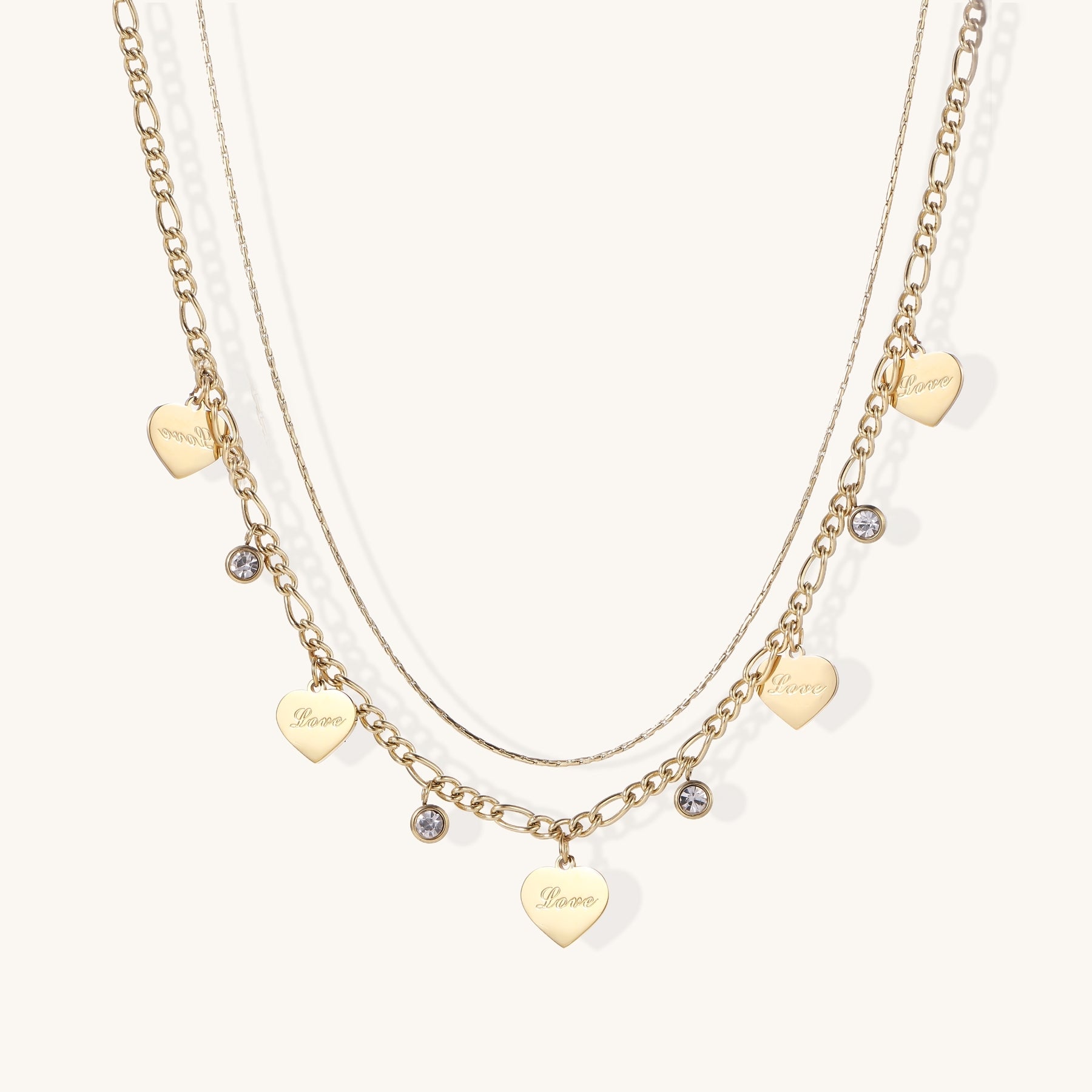 Two layered gold-plated necklaces with heart pendants engraved 'Love' and sparkling crystal charms on white background.