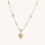 Dainty 14K gold plated necklace with heart pendant and bezel-set crystals on delicate chain against cream background.