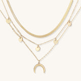 Gold-plated layered necklace set featuring herringbone chain, disc pendants, and crescent moon charm on beige background.