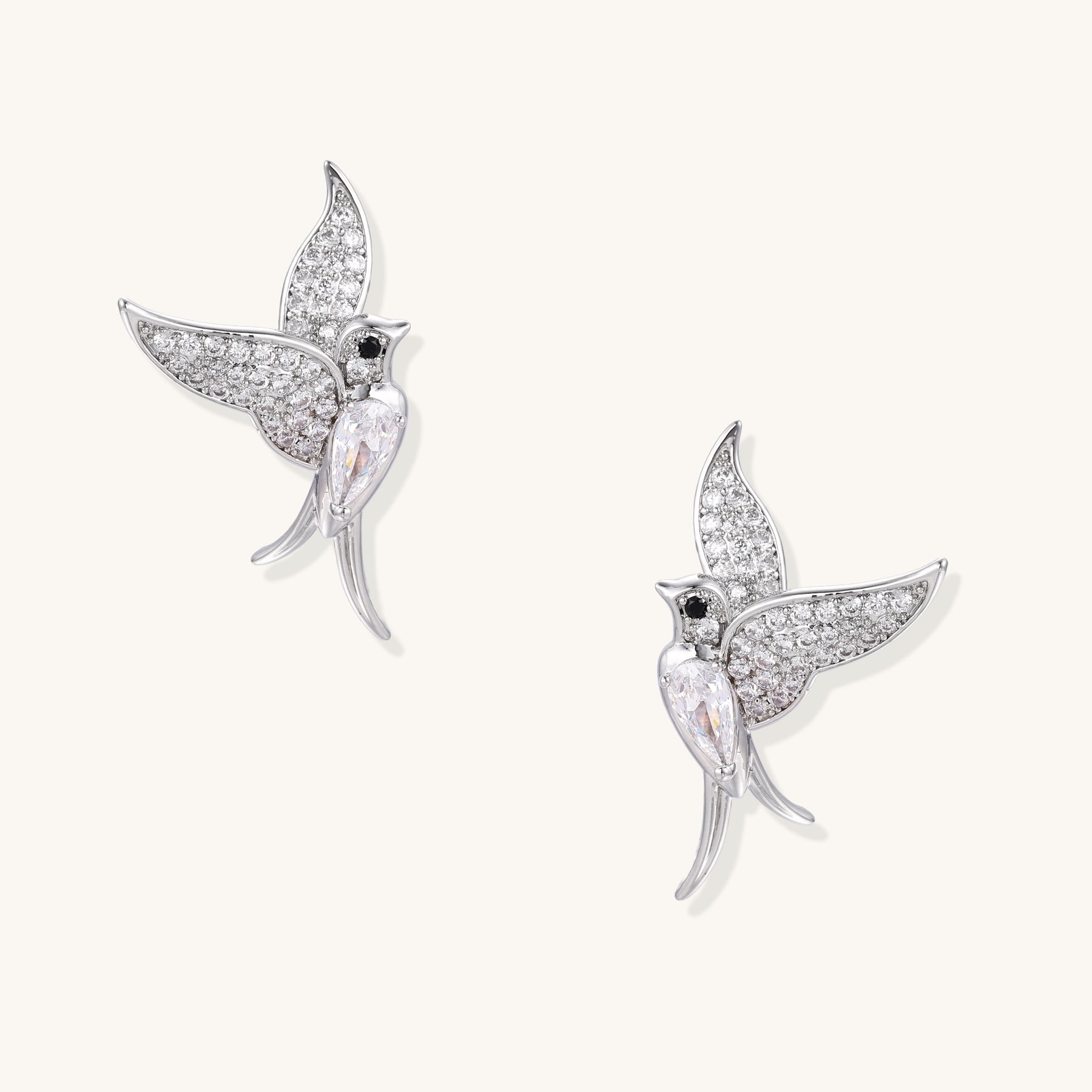 Pair of gold-plated hummingbird earrings with crystal pavé wings and black stone eyes on neutral background.