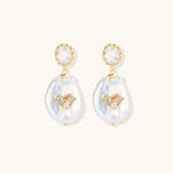 Gold-plated drop earrings with iridescent baroque pearls and crystal flower accents on cream background