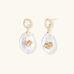 Gold-plated drop earrings with iridescent baroque pearls and crystal flower accents on cream background