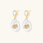 Gold-plated drop earrings with iridescent baroque pearls and crystal flower accents on cream background