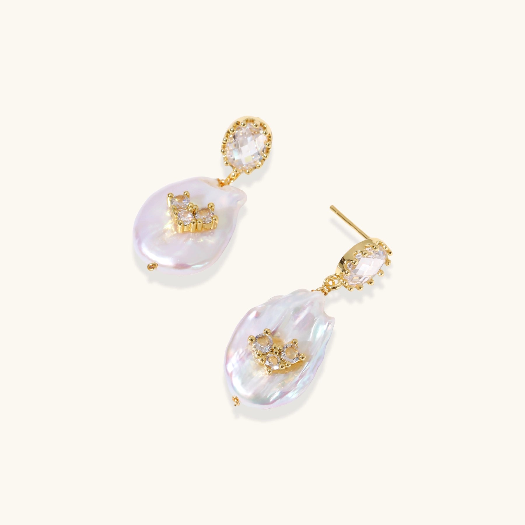 Gold-plated drop earrings with iridescent baroque pearls and crystal flower accents on cream background
