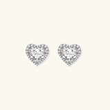 Close-up of heart-shaped moissanite stud earrings in sterling silver halo setting on cream background.