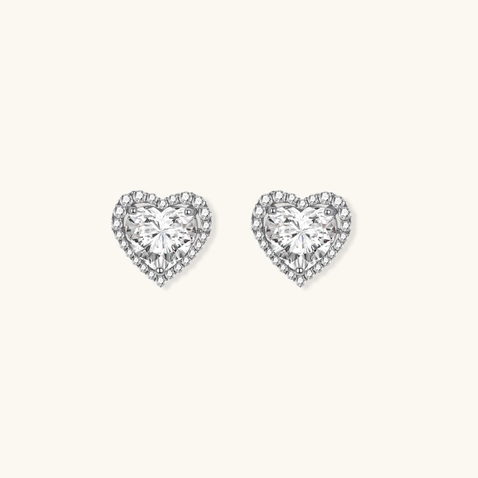 Close-up of heart-shaped moissanite stud earrings in sterling silver halo setting on cream background.