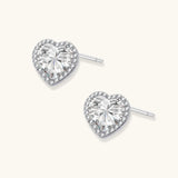 Close-up of heart-shaped moissanite stud earrings in sterling silver halo setting on cream background.