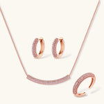 Rose gold jewelry set with pavé cubic zirconia stones including curved necklace, hoop earrings, and adjustable ring on cream background.