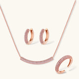 Rose gold jewelry set with pavé cubic zirconia stones including curved necklace, hoop earrings, and adjustable ring on cream background.