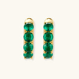 Close-up of gold-plated huggie hoop earrings featuring four emerald green cubic zirconia stones on white background.