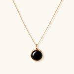 Circular black agate pendant on gold-plated sterling silver chain against cream background, showing modern minimalist design.