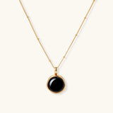 Circular black agate pendant on gold-plated sterling silver chain against cream background, showing modern minimalist design.