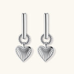 Silver huggie earrings with textured heart charms featuring radial sunburst pattern on neutral background.