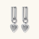 Silver huggie earrings with textured heart charms featuring radial sunburst pattern on neutral background.