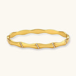 Gold-toned wavy bangle with bamboo-style segments and CZ crystal clusters on cream background. 18K plated stainless steel.
