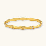 Gold-toned wavy bangle with bamboo-style segments and CZ crystal clusters on cream background. 18K plated stainless steel.