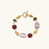 18k gold-plated copper bracelet with burgundy stones, smoky quartz, baroque pearls and ornate golden charms on white background.