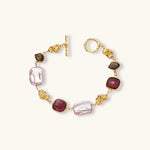 18k gold-plated copper bracelet with burgundy stones, smoky quartz, baroque pearls and ornate golden charms on white background.